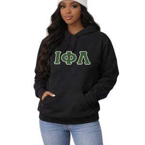 May include: A black hooded sweatshirt and white beanie with the Greek letters IΦA in green. The sweatshirt has a front pocket and the letters are also on the front. The person is wearing blue jeans.