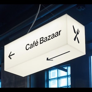 Könnte beinhalten: Ein rechteckiges, beleuchtetes Schild für "Café Bazaar" hängt von der Decke. Das weiße Schild hat schwarzen Text und Richtungspfeile sowie ein Messer- und Gabel-Symbol. Das Schild hängt vor einem dunklen Hintergrund.