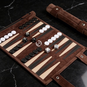 May include: A brown leather backgammon set with black and white game pieces and dice. The board is open, displaying the playing surface with alternating light and dark triangles. A matching brown leather dice holder is also present.