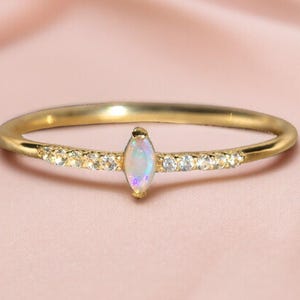 May include: A gold ring with a marquise-shaped opal centre stone. The band is embellished with small, sparkling accent stones. The ring is set against a soft pink background, emphasising its elegant design.