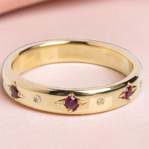 May include: A gold band ring with star-shaped settings, each holding a deep red gemstone. Small, clear gemstones are interspersed between the stars. The ring is set against a soft pink background.