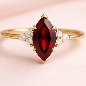 May include: A gold ring featuring a marquise-cut, deep red gemstone as the centerpiece. The ring is accented with small, round, white gemstones on either side of the main stone. The band is a thin, polished gold.