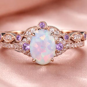 May include: A rose gold ring set featuring an oval opal center stone. The ring is accented with small purple gemstones and clear crystals. The band is also adorned with small crystals. The ring is set on a pink satin surface.