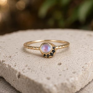 Puede incluir: Un anillo de oro con un cabujón central redondo de piedra lunar iridiscente, acentuado por un grupo de pequeñas piedras negras en forma de media luna en un lado. La banda del anillo tiene un patrón texturizado de estilo trenzado.