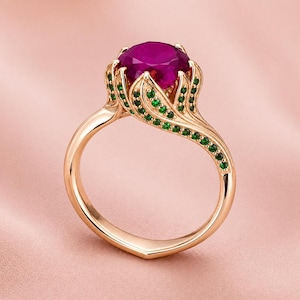 May include: A gold ring featuring a large, round, magenta gemstone. The ring's setting has a floral design with small green gemstones. The band is a smooth, polished gold. The ring is set against a soft pink background.