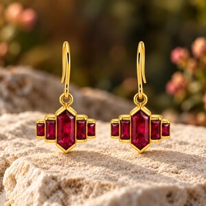 May include: A pair of gold drop earrings featuring a central hexagonal red gemstone flanked by two smaller rectangular red gemstones on each side. The earrings hang from simple gold hooks, showcasing a symmetrical, geometric design.