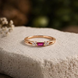 May include: A gold ring featuring a central rectangular baguette-cut pink gemstone flanked by three small round clear stones on each side. The band is thin and polished, showcasing a delicate design that highlights the vibrant pink center stone.