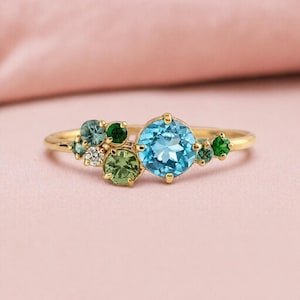 Può includere: Un anello d'oro con un gruppo di pietre preziose. L'anello presenta una grande pietra preziosa rotonda blu chiaro, circondata da pietre preziose verdi e bianche più piccole. La fascia è in oro sottile e lucido. Diametro approssimativo: 1,9 cm.