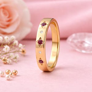 May include: A gold ring with a flat band, featuring three red gemstone accents in star-shaped settings, and three small clear gemstones. The ring is displayed on a pink surface, with a rose and small white flowers in the background.