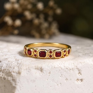 Pode incluir: Um anel de banda dourada com um padrão repetitivo de gemas vermelho-escuras, alternando cortes octogonais e pequenos cortes redondos. O metal tem um acabamento amarelo polido e as pedras estão engastadas de forma segura no anel.