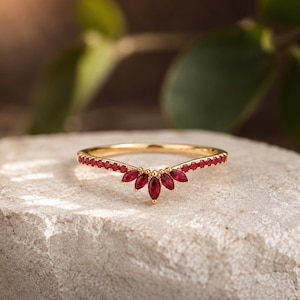 May include: A gold chevron-shaped ring featuring a row of small round red gemstones along the band and a cluster of five marquise-cut red gemstones arranged in a fan pattern at the center point of the V-shaped design.