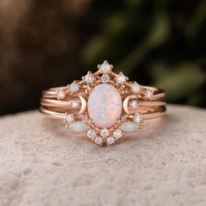 May include: A rose gold ring set featuring a central oval opal cabochon flanked by two crescent moon shapes. The design is accented with small round clear stones and two marquise-cut opals, arranged in a symmetrical, tiered crown-like pattern.