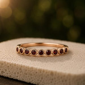 Op de afbeelding: Een gouden ring met een rij kleine, ronde, dieprode granaat edelstenen. Elke edelsteen is gezet in een cirkelvormige, kralen metalen zetting, wat zorgt voor een doorlopend, gestructureerd ontwerp langs de bovenste helft van de gepolijste gouden ring.