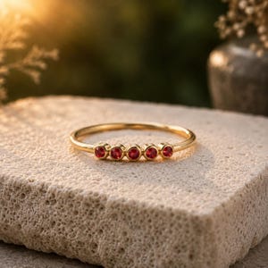 Può includere: Un sottile anello in oro con cinque piccole pietre preziose rotonde di un rosso vivace, disposte in fila sulla parte superiore della fascia. Il metallo ha una finitura lucida e le pietre sono fissate in castoni circolari individuali.