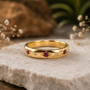 May include: A gold band ring featuring a repeating pattern of round red rubies and smaller clear white diamonds. The gemstones are set into the polished gold surface, with the rubies framed by four-pointed star-shaped engravings in the metal.