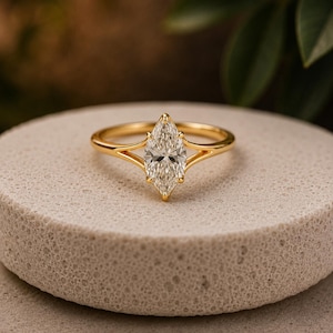 May include: A gold ring featuring a marquise-cut clear gemstone held by a six-prong setting. The band has a split shank design that tapers toward the center stone, creating a sleek and polished appearance with a bright yellow metallic finish.