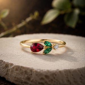 May include: A gold ring featuring a pear-shaped red gemstone, two marquise-cut green gemstones arranged like leaves, and a single small round clear gemstone set on a thin polished gold band, creating a floral-inspired cluster design.