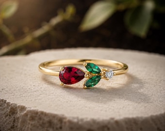 Op de afbeelding: Een gouden ring met een peervormige rode edelsteen, twee marquise-geslepen groene edelstenen in de vorm van blaadjes en een kleine ronde heldere edelsteen op een dunne gepolijste gouden band, wat een bloemachtig ontwerp creëert.