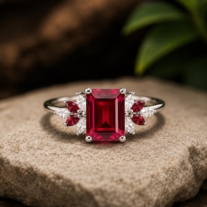 May include: A silver-toned ring featuring a large emerald-cut red gemstone at the centre. The setting is accented on both sides by a cluster of smaller marquise-cut red and clear white gemstones arranged in a symmetrical, leaf-like pattern.