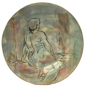 May include: A circular ceramic plate with a hand-painted design of a seated figure, seen from behind. The figure is depicted in black line art, set against a backdrop of soft greens, browns, and pinks. The plate's rim has a gentle, natural colour.