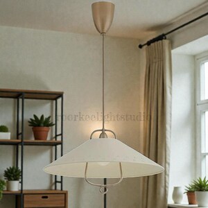 May include: Cream-coloured pendant light with a conical shade and a metal frame. The light fixture hangs from a thin cord and has a metallic finish. The design is simple and modern, suitable for various interior styles.