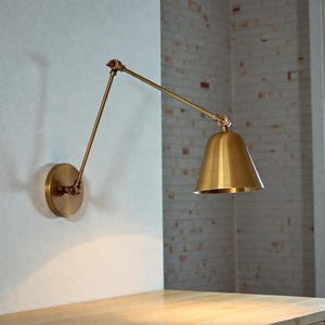 Könnte beinhalten: Eine Wandleuchte aus Messing mit verstellbarem Arm und konischem Schirm. Die Lampe ist an einer runden Basis an der Wand befestigt. Das Design ist minimalistisch und industriell, passend für verschiedene Einrichtungsstile.