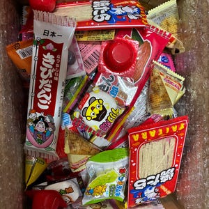 Peut inclure: Une bo&icirc;te en carton remplie de diverses collations japonaises. L'assortiment comprend des bonbons emball&eacute;s individuellement, des collations de poisson s&eacute;ch&eacute; et d'autres friandises. Les couleurs des emballages comprennent le rouge, le blanc, le jaune et le vert. Certains emballages comportent du texte japonais et des illustrations de dessins anim&eacute;s.
