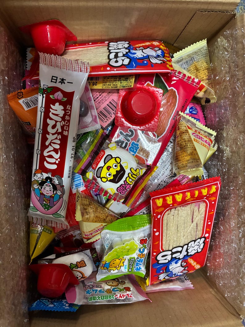 Peut inclure: Une bo&icirc;te en carton remplie de diverses collations japonaises. L'assortiment comprend des bonbons emball&eacute;s individuellement, des collations de poisson s&eacute;ch&eacute; et d'autres friandises. Les couleurs des emballages comprennent le rouge, le blanc, le jaune et le vert. Certains emballages comportent du texte japonais et des illustrations de dessins anim&eacute;s.