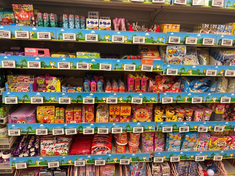 Peut inclure: &Eacute;tag&egrave;re de supermarch&eacute; remplie de diverses collations et bonbons japonais. Les &eacute;tag&egrave;res sont d&eacute;cor&eacute;es d'un motif tropical bleu et vert. Les produits comprennent des bonbons emball&eacute;s, des gommes et d'autres friandises.