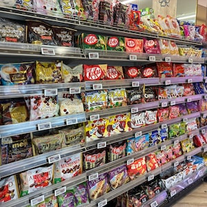 Peut inclure: Rayonnage de supermarch&eacute; rempli de divers snacks emball&eacute;s. Les &eacute;tag&egrave;res sont garnies de sacs et de bo&icirc;tes color&eacute;s de bonbons, chocolats et biscuits. Des marques comme Kit Kat et Meiji sont visibles, offrant un large choix.