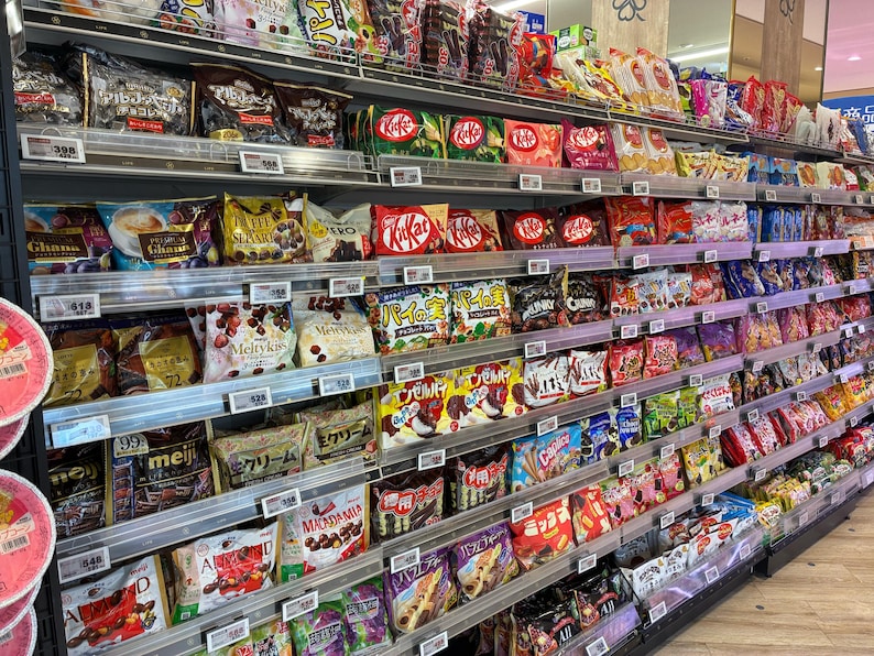 Peut inclure: Rayonnage de supermarch&eacute; rempli de divers snacks emball&eacute;s. Les &eacute;tag&egrave;res sont garnies de sacs et de bo&icirc;tes color&eacute;s de bonbons, chocolats et biscuits. Des marques comme Kit Kat et Meiji sont visibles, offrant un large choix.