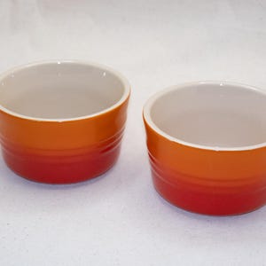 Op de afbeelding: Twee kleine, ronde ramekins met een kleurenschema. De buitenkant loopt over van een dieprode kleur aan de basis naar een feloranje aan de bovenkant. De binnenkant is lichtcrème van kleur. Deze zijn waarschijnlijk voor het bakken of serveren van individuele porties.