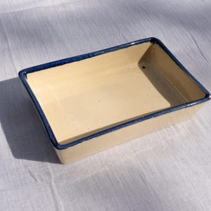 Könnte beinhalten: Eine rechteckige Keramik-Backform mit cremefarbenem Innenraum und dunkelblauem Rand. Die leere Form liegt auf einem weißen Tuch, mit einem Schatten zur Seite. Die Form wird wahrscheinlich zum Backen oder Servieren von Speisen verwendet.