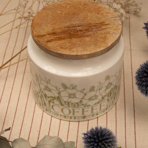 Può includere: Un contenitore per caffè in ceramica bianca con un motivo floreale verde chiaro attorno al centro e la parola COFFEE stampata in lettere maiuscole. Il contenitore è sormontato da un coperchio rotondo in legno naturale con venature visibili.