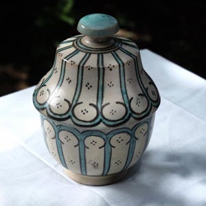 Può includere: Un barattolo in ceramica con coperchio caratterizzato da una base color crema decorata con strisce verticali color verde acqua e contorni neri. Il motivo è composto da forme smerlate ripetitive e piccoli gruppi di punti.