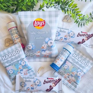 Peut inclure: Divers cadeaux de baby shower, comprenant un paquet de chips Lay's, des barres chocolatées Kinder Bueno, des bonbons Haribo et des bulles de savon. Les articles sont décorés de canards et du texte "Baby Shower de Caroline".