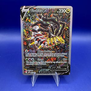 May include: A Giratina V Pokémon trading card. The card features a black and white creature with yellow accents, set against a colorful, detailed background. The card displays the text "Abyss Seeking" and "Shred", along with other game-related information.