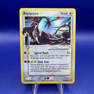May include: A Rayquaza Pokémon trading card with a yellow border. The card features a black and teal illustration of Rayquaza. Text includes "Rayquaza", "90 HP", and attack descriptions. The card is displayed on a clear plastic stand.