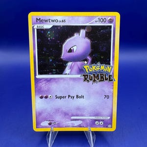 May include: A Pokémon Rumble trading card featuring Mewtwo Lv.65. The card has a purple and yellow border with the text "Super Psy Bolt" and "70". The card also has the Pokémon Rumble logo and the HP is 100.