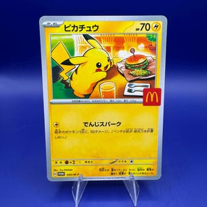 Könnte beinhalten: Eine Pokémon-Sammelkarte mit Pikachu, einer Abbildung von Burger und Getränk. Die Karte hat japanischen Text und ein McDonald's-Logo. Die Karte wird auf einem transparenten Kunststoffständer vor blauem Hintergrund präsentiert.