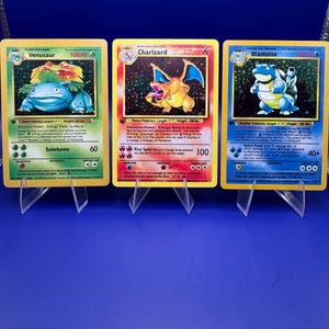 Op de afbeelding: Drie Pokémon-verzamelkaarten op transparante standaards. De kaarten tonen Venusaur, Charizard en Blastoise, elk met unieke illustraties en statistieken. De kaarten zijn in goede staat, met levendige kleuren en duidelijke tekst.