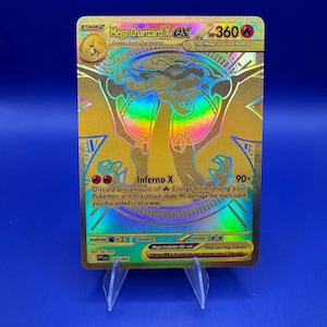 Könnte beinhalten: Eine goldene Mega Charizard X EX Pokémon-Karte. Die Karte zeigt ein holografisches Bild von Charizard mit dem Text "Inferno X" und "360 HP". Die Karte wird auf einem transparenten Ständer vor blauem Hintergrund präsentiert.