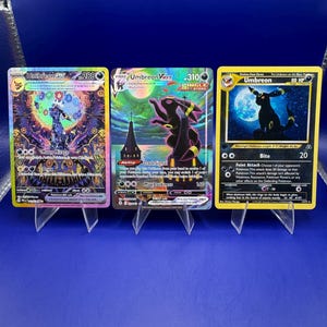 May include: Three Umbreon Pokémon trading cards displayed on clear stands. The cards feature Umbreon in various designs, including a VMAX card with a dark silhouette and a classic card with a moonlit background. The cards are set against a blue backdrop.