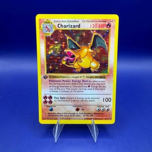 May include: A Charizard Pokémon trading card, Stage 2, with a yellow border. The card features a fiery orange dragon with green wings. Text includes "Charizard", "120 HP", and attack details. The card is displayed on a clear plastic stand.