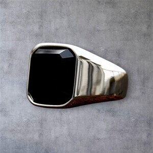 May include: A silver-toned signet ring featuring a large, rectangular, faceted black stone set into the center. The band has a smooth, polished metallic finish that reflects light, creating a clean and solid appearance for this piece of jewelry.