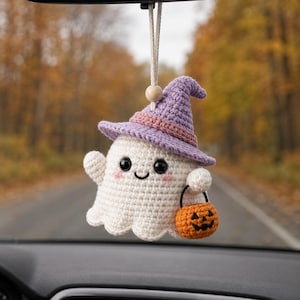 May include: A crocheted ghost ornament featuring a light purple witch hat with a pink band, black safety eyes, pink cheeks, and a small orange jack-o'-lantern bucket. The ghost hangs from a white cord with a single wooden bead at the top.