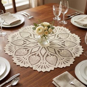 May include: A round, cream-coloured crochet doily featuring an intricate, repeating pineapple-inspired pattern with a scalloped edge. The doily is centred on a wooden table, serving as a base for a clear glass vase containing white flowers.