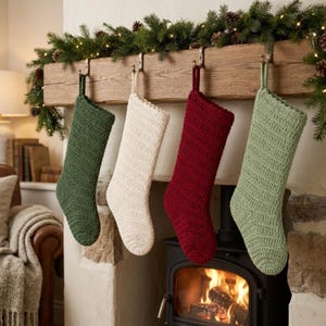 May include: Four crocheted Christmas stockings hanging from a wooden mantel. The stockings are solid colours: dark green, cream, deep red, and sage green. Each stocking features a textured, ribbed stitch pattern and a loop at the top for hanging.