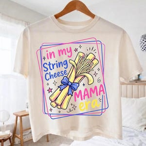 以下が含まれることがあります： クリーム色のTシャツ。青いリボンで結ばれたストリングチーズのグラフィックと、カラフルなフレームに「in my String Cheese MAMA era」というテキストがプリントされています。デザインには星や装飾的な要素が含まれています。