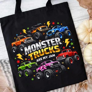 May include: Black tote bag featuring a colorful graphic of monster trucks. The design includes various trucks in orange, blue, green, pink, and red, with the text "MONSTER TRUCKS ARE MY JAM" and lightning bolt accents.
